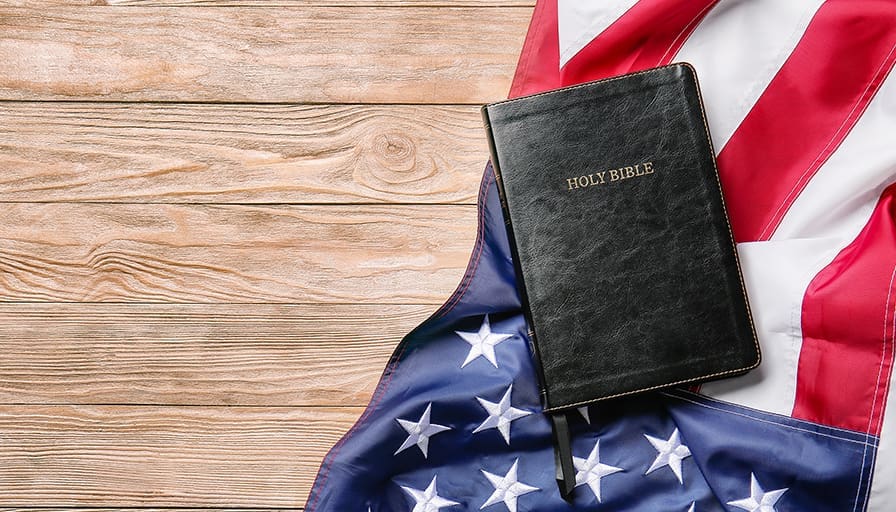 A black leather Holy Bible resting on an American flag spread across a wooden surface.